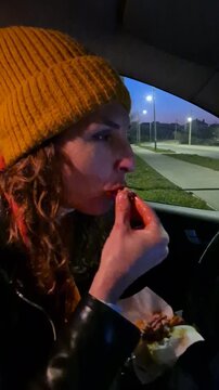 Woman eating food inside car at night near illuminated suburban street while a pedestrian walks past. Close-up static camera shot.