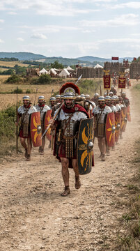 Una legi&oacute;n de soldados romanos con armadura, escudos y estandartes marcha en formaci&oacute;n por un camino de tierra junto a un fuerte de madera.