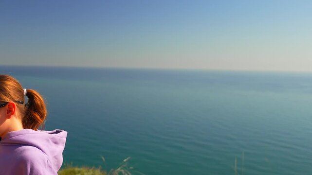 Coastal cliffside setting features a young girl wearing sunglasses dancing energetically against the blue sea. Panning and tilting camera shot.
