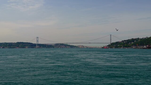 A wide landscape shot of the iconic suspension bridge crossing the Bosphorus strait in Istanbul with calm blue waters and distant hills under an overcast sky