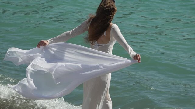 Ocean waves crash against a woman in a sheer gown as she holds the fabric outdoors. Static medium shot.