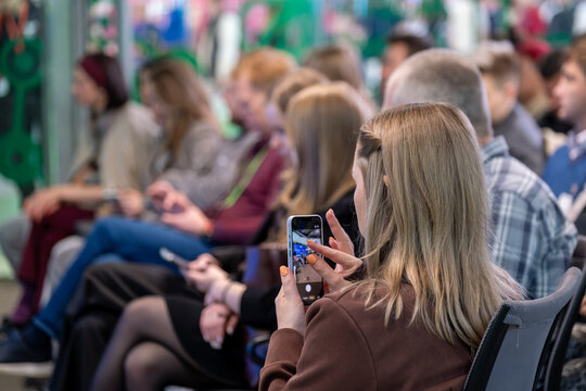 Woman in conference audience photographs speaker with smartphone, attendees sit in rows and use devices.