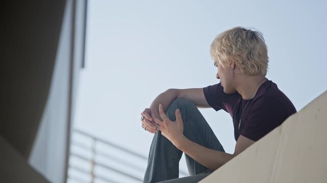 Caucasian teenager sitting outdoors on concrete ledge, looking away and fidgeting with rings. Footage is ideal for youth mental health campaigns or social issue videos