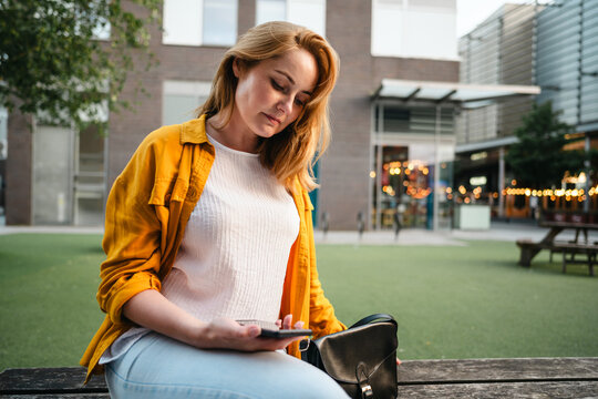 A woman sits on a bench in the city and texts on her smartphone