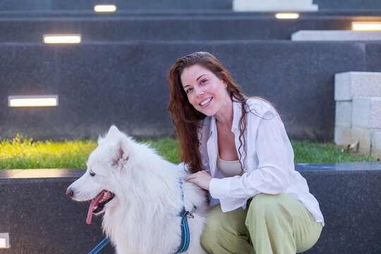 A smiling woman pets her white samoyed dog outdoors on stone steps