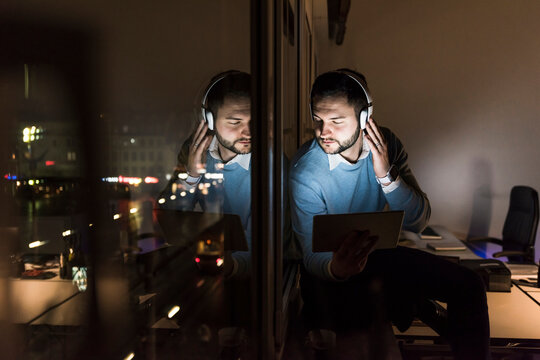 Businessman sitting on window sill in office at night listening music with headphones
