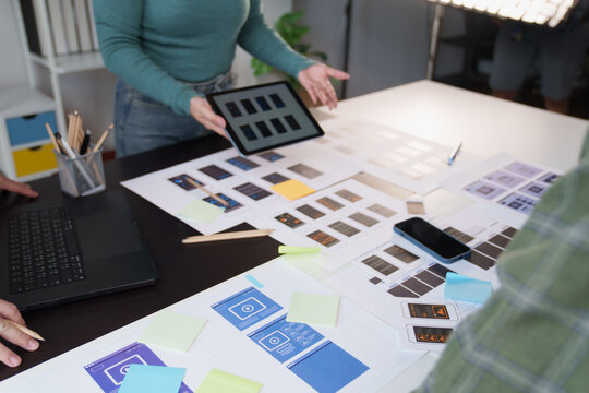 UX designers collaborate over mobile app wireframes and prototypes on a desk, reviewing screen layouts on tablet and laptop to plan, iterate and refine the user experience