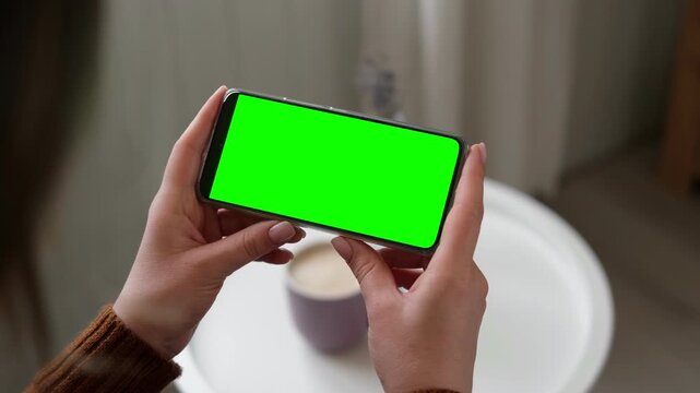 Girl sitting at home and watching movie on mobile phone template mockup with green screen chroma key. Female hands holding smartphone horizontally for digital entertainment leisure in cozy room