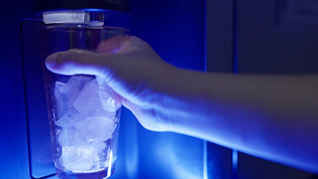 Close up a hand positions a glass filled with ice cubes under a refrigerator's water dispenser with vibrant blue LED light shines