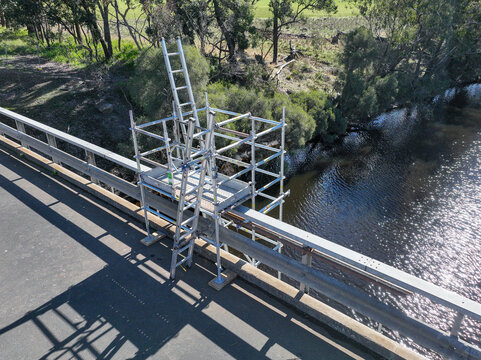 Scaffolding tower installed on a rural bridge to provide safe access for maintenance and inspection work. Metal platforms and supports surround the bridge structure over a small waterway.
