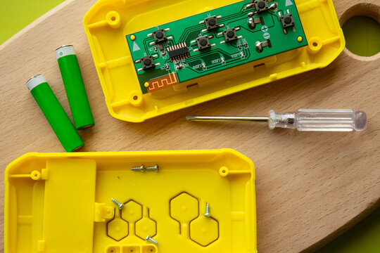Disassembled electronic device with green circuit board, yellow plastic case and AA batteries. Top view flat lay of PCB motherboard hardware components repair process.