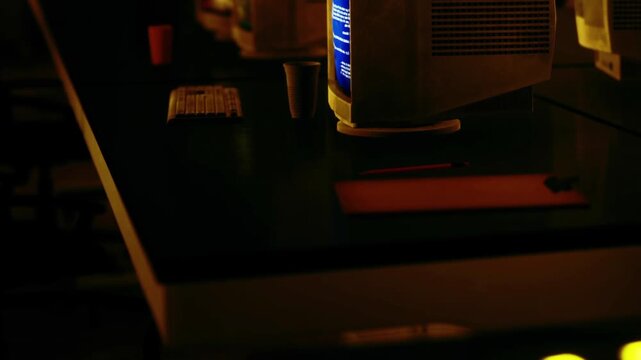 A room filled with old computers sits under low light. The screens glow softly while various items are placed on the desks. It is night and the atmosphere feels quiet.