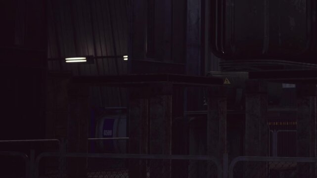 A dark industrial setting at night. Machinery and structures are present with artificial lighting. The atmosphere feels heavy and work focused.