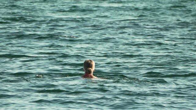 Woman swimming breaststroke through calm ocean waves under bright daylight. Static medium telephoto shot.