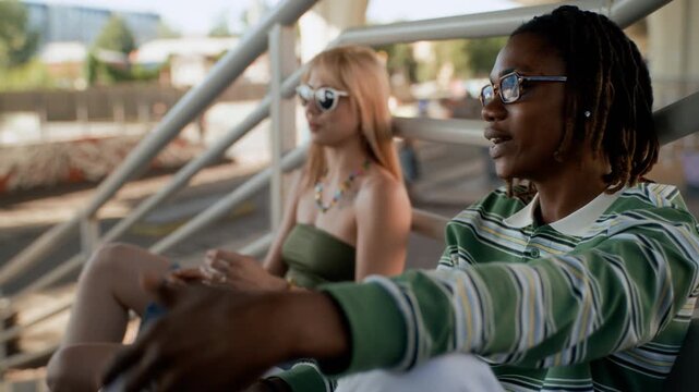 African American young adult talking and gesturing while sitting on outdoor stairs next to Caucasian young woman. Clip is ideal for youth lifestyle and diversity marketing campaigns