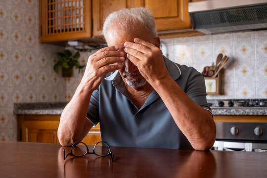 Senior man sitting at a kitchen table, experiencing a painful headache and fatigue, rubbing his temples and covering his eyes due to stress and visual discomfort