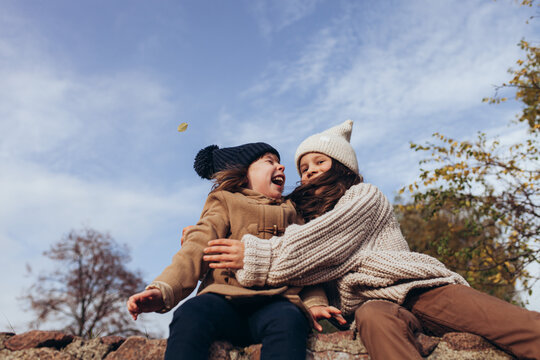 Two happy little girls in warm autumn clothes laughing and hugging outdoors against blue sky. Childhood, friendship, joy, warmth, family bonding, carefree play.