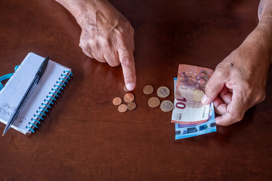 Elderly person's hands counting euro coins and banknotes on a wooden table, managing a budget with a notebook, symbolizing retirement planning, financial challenges, and economic hardship