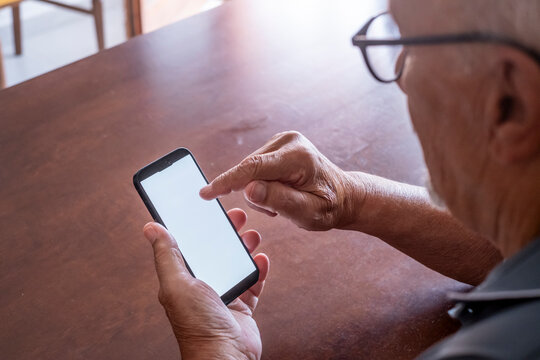 Senior man with glasses using a smartphone with a blank white touch screen, interacting with digital technology and communication while sitting at a wooden table