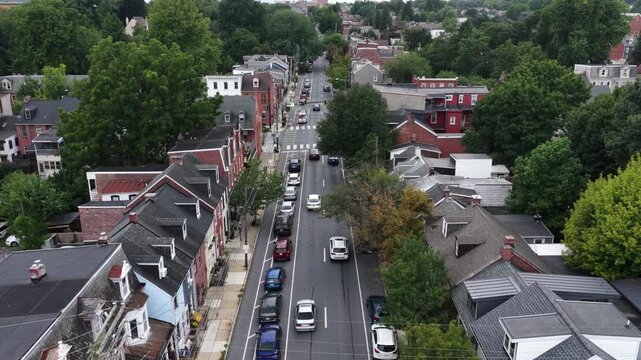 Traffic moves past brick row houses on residential street in Lancaster, Pennsylvania, parked cars and shaded sidewalks capture classic small town American housing charm. Aerial drone rising shot
