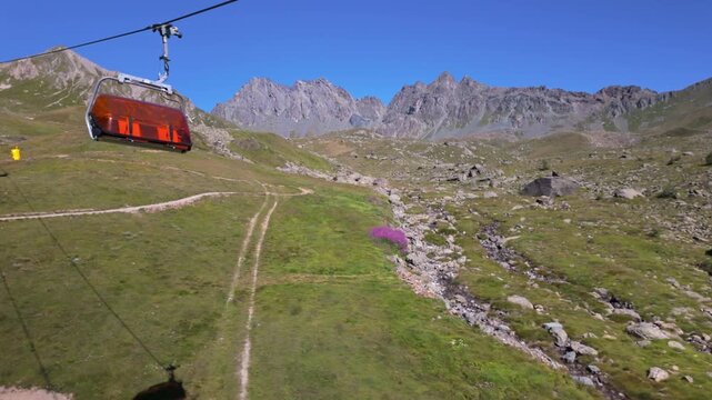 Rocky mountain ridge above green alpine meadows, viewed from a cable car with a winding trail, small stream and clear blue summer sky.