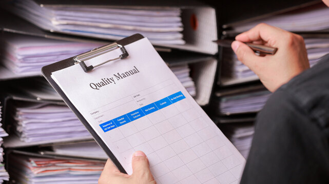 Focused auditor conduct detailed review on document attached to clipboard inside busy office filled with file and binder during audit