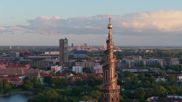 Drone orbit around the belfry of Our Saviour's Church Belfry (Vor Frelsers Kirke), revealing Copenhagen's cityscape bathed in warm sunset light and golden-hour tones.