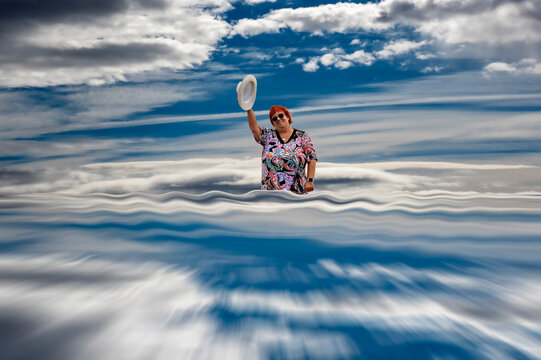 Persona con camisa colorida, gafas y sombrero blanco alzado; cielo dram&aacute;tico y reflejo acu&aacute;tico que duplica la escena; composici&oacute;n surreal, sim&eacute;trica y llena de movimiento. 