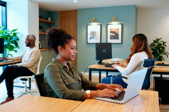 Black young adult woman working on laptop in modern office while colleagues using desktop computers, representing teamwork and productivity. Ideal for business, staffing, workplace content