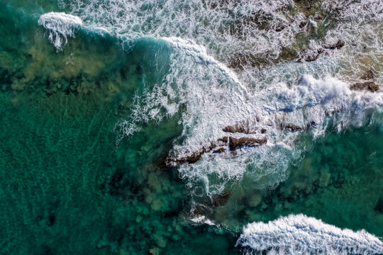 Aerial downward view of ocean waves crashing over rugged rock formations at Lake Arragan Beach, capturing white surf, exposed coastal geology and the powerful movement of the Pacific Ocean 