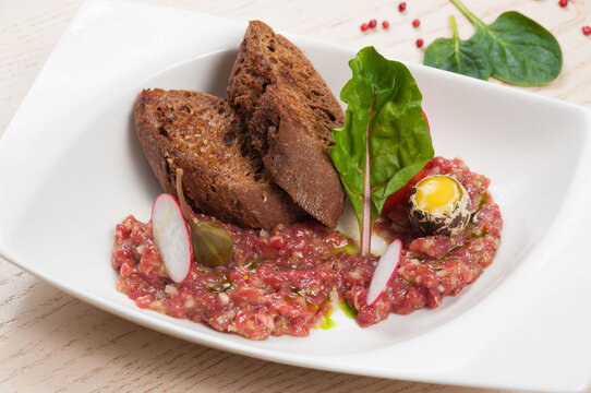 Beef steak tartare with quail egg yolk, toasted rye bread, and radish on white plate