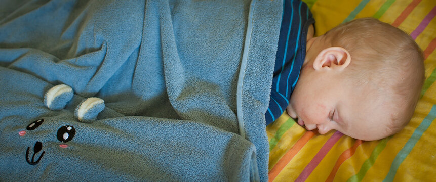 A peaceful baby boy sleeps soundly in a bear-patterned blue blanket on colorful striped sheets