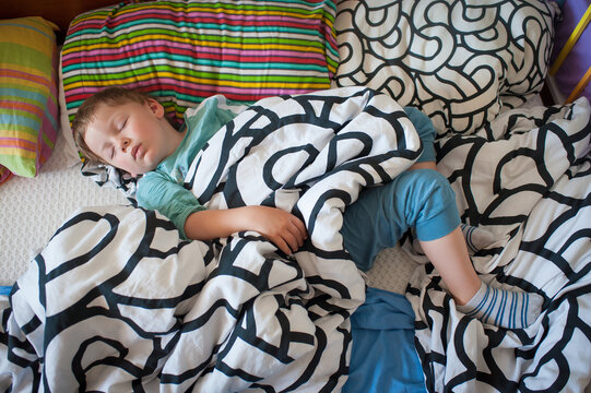 Peaceful young boy curled up in soft blankets sleeping soundly during a quiet afternoon nap in his cozy bed surrounded by colorful patterned pillows