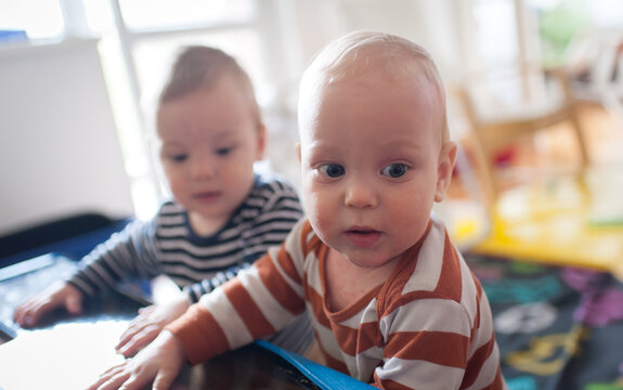 Two curious twin babies exploring their home nursery during a bright morning while reaching out to touch a smooth dark surface with wide excited eyes