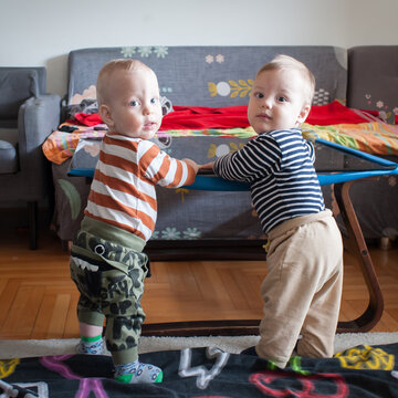 Twin brothers in colorful outfits stand tall, holding a glass table in their cozy living room while looking at the camera