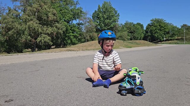 A cheerful toddler in a striped tee and navy shorts sits on sunlit asphalt, legs stretched slightly and hands resting by his side; a teal‑and‑green rollerblade sits beside him, while leafy trees and a