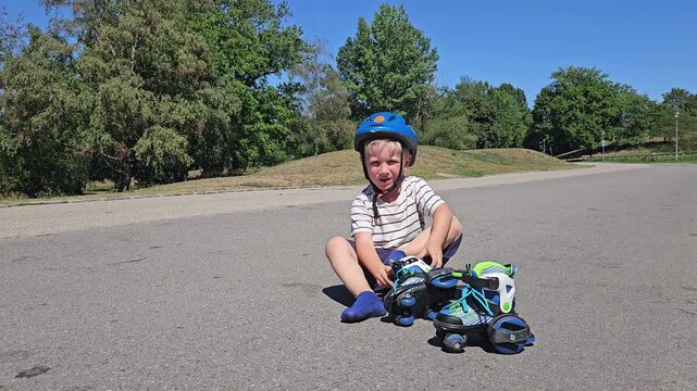 A smiling toddler in a striped tee and a vivid blue safety helmet sits on sunlit asphalt, legs bent and hands resting on knees; a teal‑and‑green rollerblade lies nearby, while verdant trees and a clea