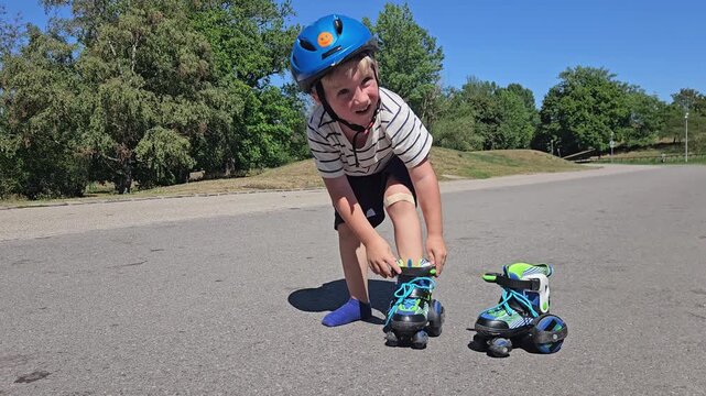 A boy in a striped shirt and a colorful safety helmet bends to adjust his rollerblade on a sunlit asphalt road; the second rollerblade rests nearby, while leafy trees and a clear sky complete the summ