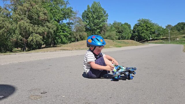 A boy in a striped tee and a vivid blue safety helmet checks his rollerblade while seated on a quiet asphalt road; verdant trees and a bright sky create a relaxed, natural backdrop