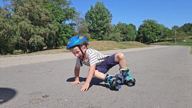 A young boy in a striped shirt and a bright blue safety helmet sits on a sunlit asphalt road after a fall, one rollerblade still on his foot; lush green trees and a clear blue sky frame the candid mom