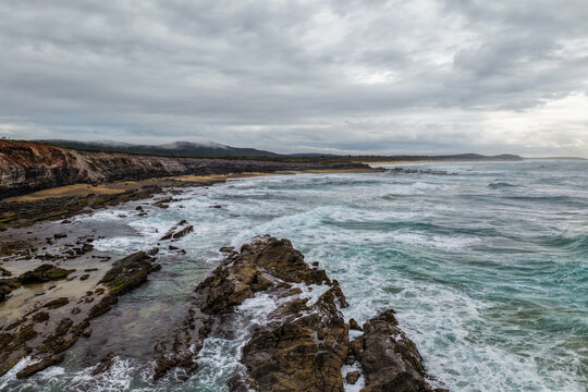 Aerial view of ocean waves crashing onto the rugged rocky coastline at Lake Arragan Beach beneath a cloudy sky, showcasing dramatic surf, exposed rock formations and the wild Pacific coastline