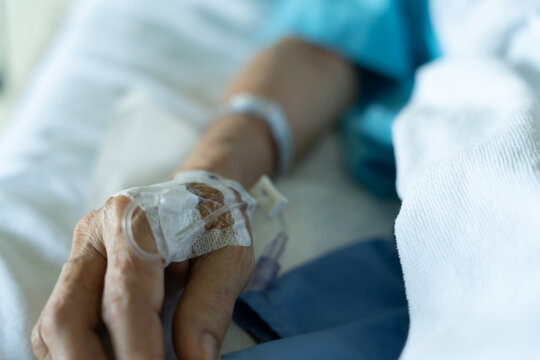 A close-up of a saline bottle strapped to a needle pricked at the hand of a female patient lying in a hospital bed. Medical and treatment of patients concepts