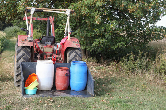 Red tractor transporting plastic barrels with water on a dry field. Concept image for water shortage, heatwave, and agricultural drought.