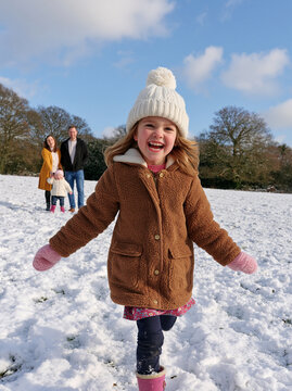 Cute little girl in teddy coat smiling and running in snow with family walking in background