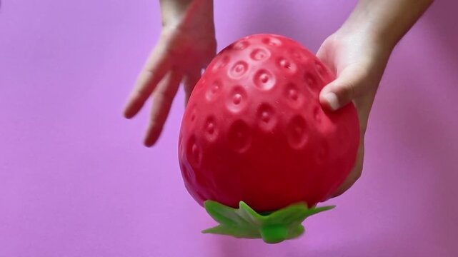 Child's hands squeezing a red strawberry shaped squishy ball for stress relief and sensory play, providing a calming and satisfying fidgeting experience against a purple background