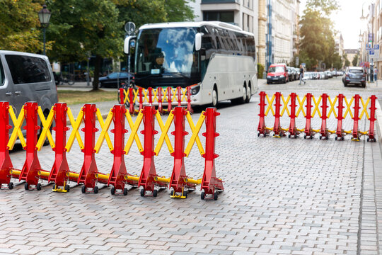 Expandable red and yellow security barriers block cobblestone street while coach waits behind temporary checkpoint. Urban traffic control creates protected route during public event in Magdeburg