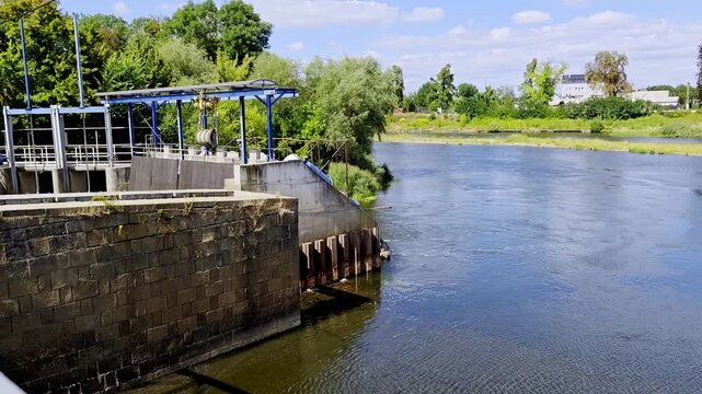 Water weir and hydrotechnical infrastructure with old stone wall on river, blue metal construction, green trees along banks and light clouds in sky on sunny day.
