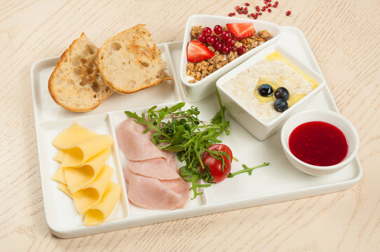 Healthy continental breakfast platter with oatmeal porridge, granola with berries, ham, cheese, and toasted bread on partitioned tray