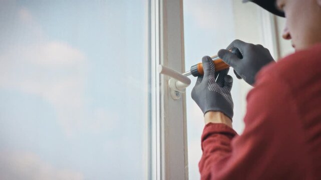 A technician in work uniform and gloves removes a plastic window handle using an orange screwdriver during a professional home repair service call. Concept of qualified window maintenance and repair