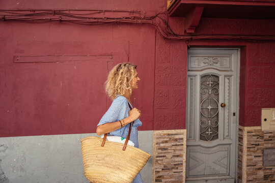Happy Woman Walking with Oversized Straw Bag Past Red Facade and Vintage Door in Southern Europe